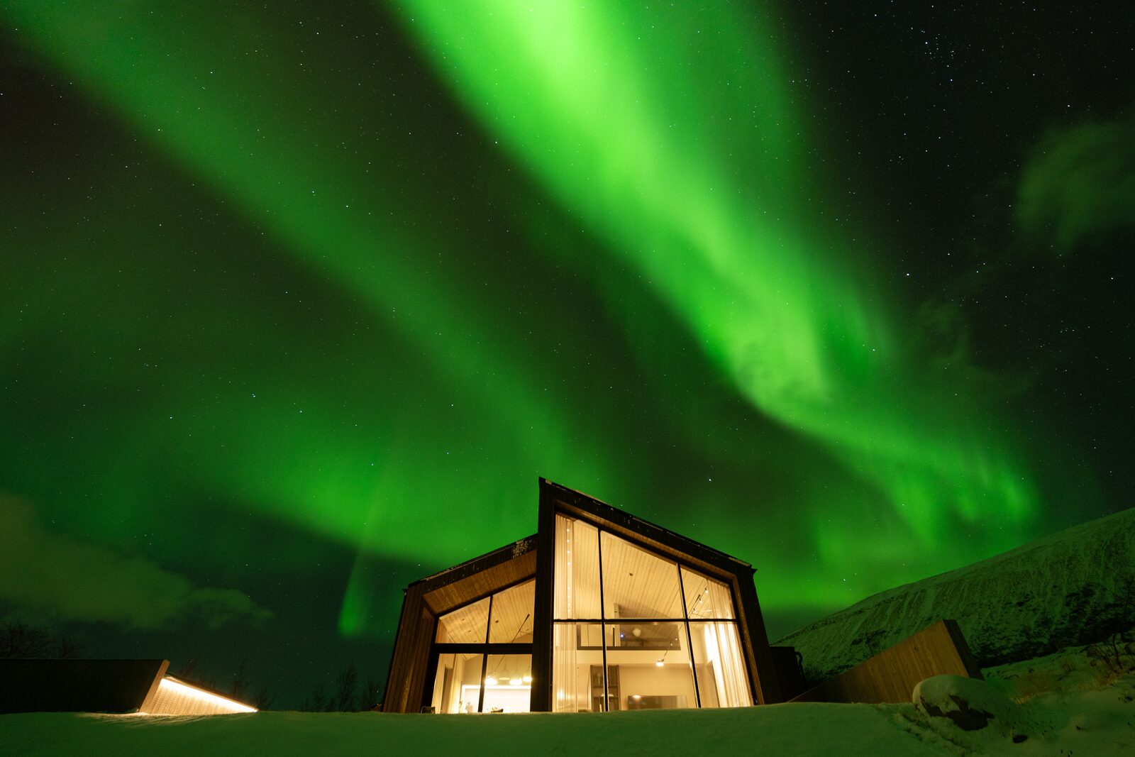 The house at Lundskógur lit from within beneath green northern lights on a snowy night.