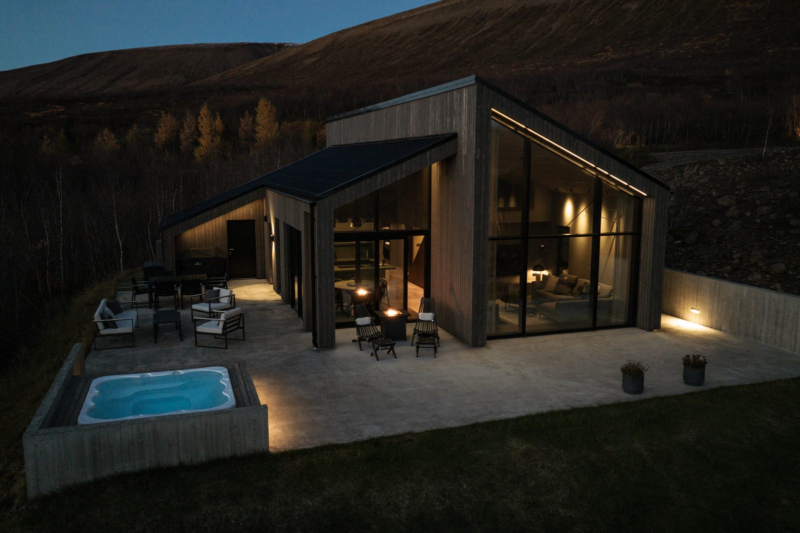 An aerial view of the house at Lundskógur at dusk, with the windows glowing, the terrace lit and the hot tub lit blue.