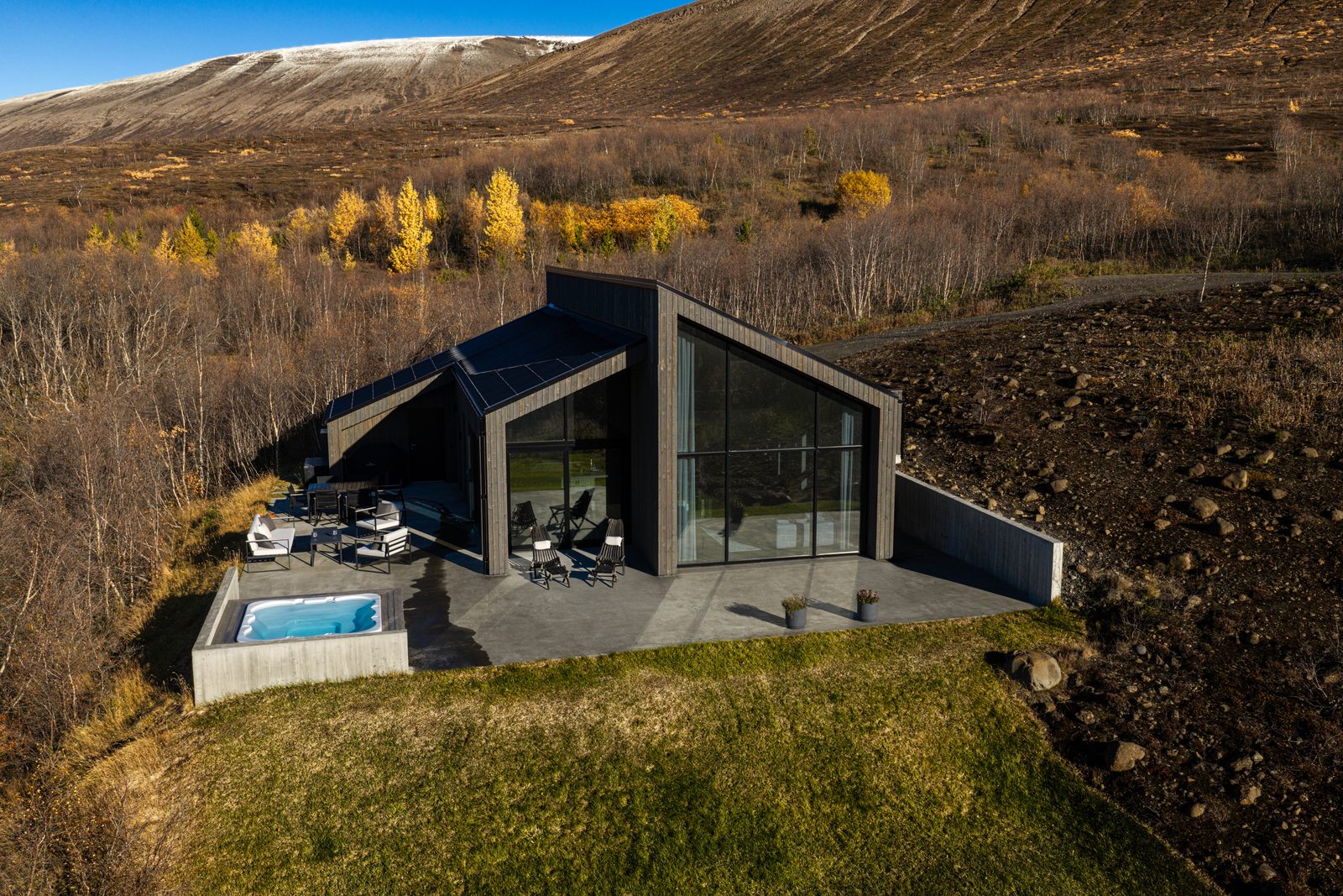 An aerial view of the house at Lundskógur with its terrace and hot tub, autumn birch forest around it and a snow-dusted fell behind.