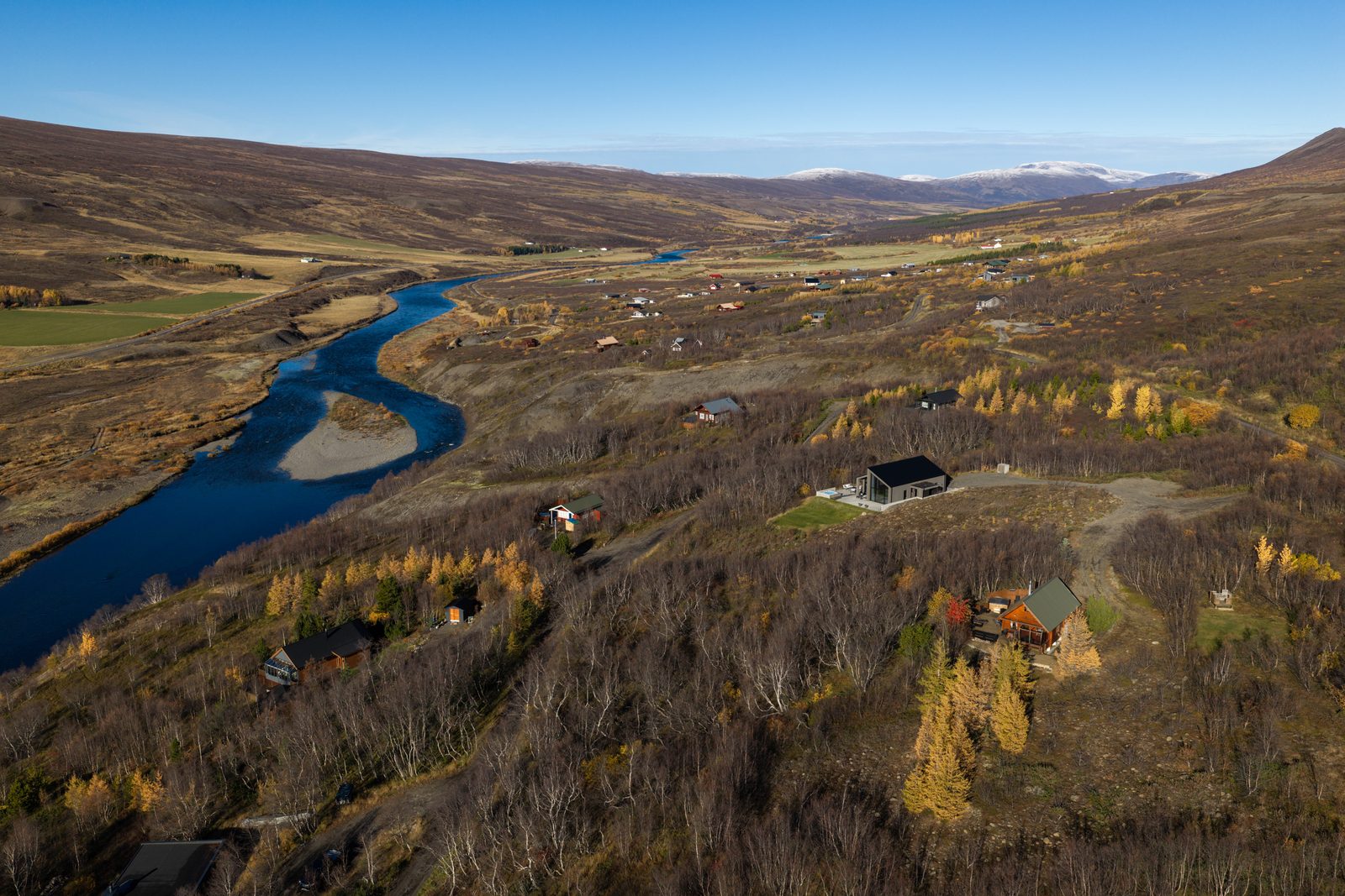 An aerial view down Fnjóskadalur in autumn, the river winding between birch forest and fields with a snow-dusted ridge beyond and the house at Lundskógur small in the frame.
