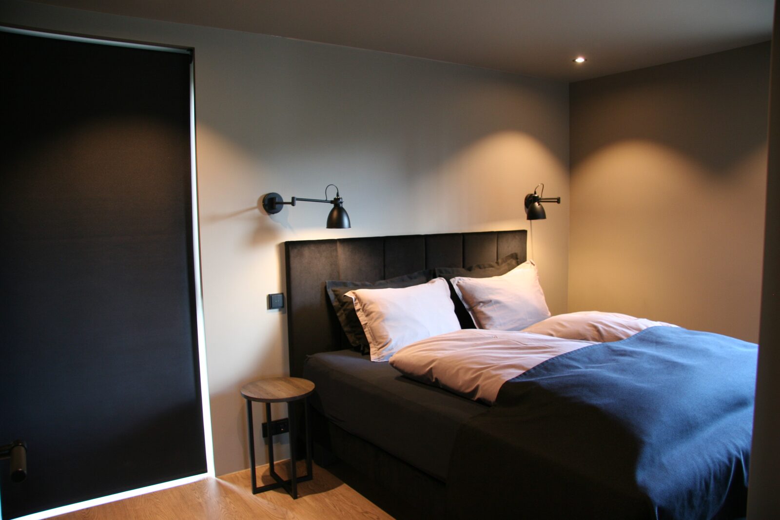 A downstairs bedroom with a queen-size bed, a dark headboard, wall lights and a window blind.