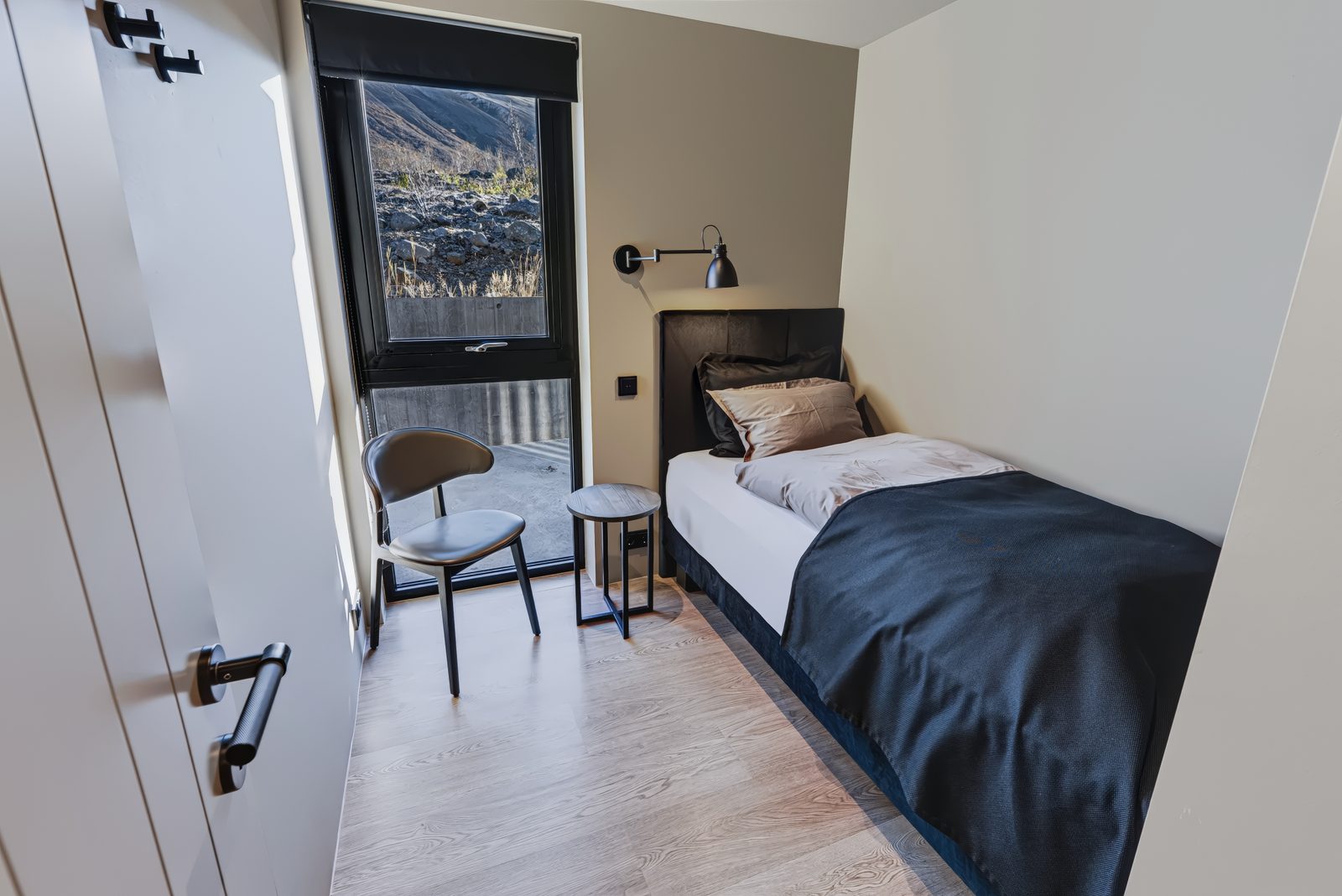 Bedroom 4 on the ground floor at Lundskógur, a single bed with a navy blanket and a dark headboard, a black wall light, a round side table and a chair beside the window.