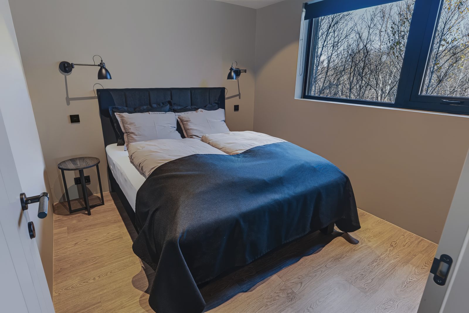 Bedroom 3 on the ground floor at Lundskógur, a queen-size bed with a black headboard and a navy blanket, a wall light on each side and a window looking out on bare birch trees.