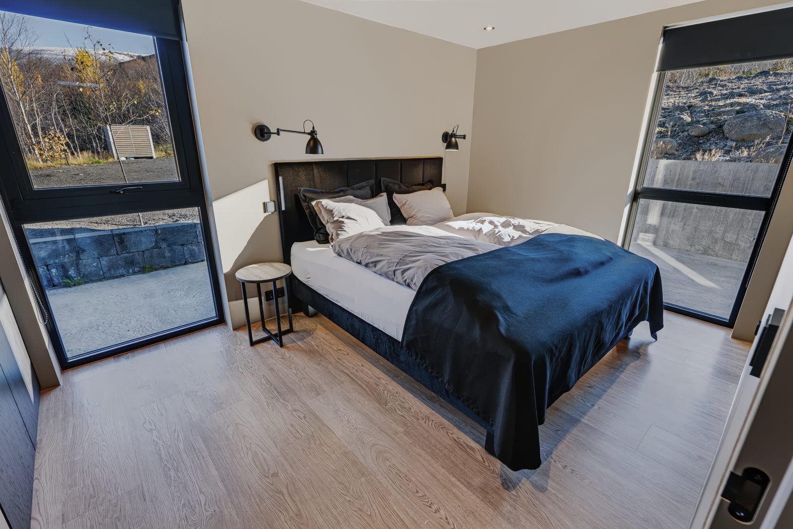 Bedroom 2 on the ground floor at Lundskógur, a queen-size bed with a navy blanket and wall lights, and two large windows with black blinds looking out on birch trees and rock.
