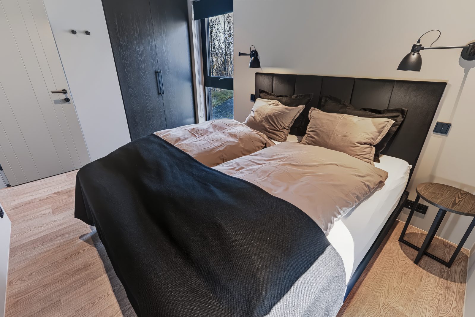 Bedroom 2 on the ground floor at Lundskógur, a queen-size bed with a black headboard and tan pillows, with a black wardrobe beside the white door.