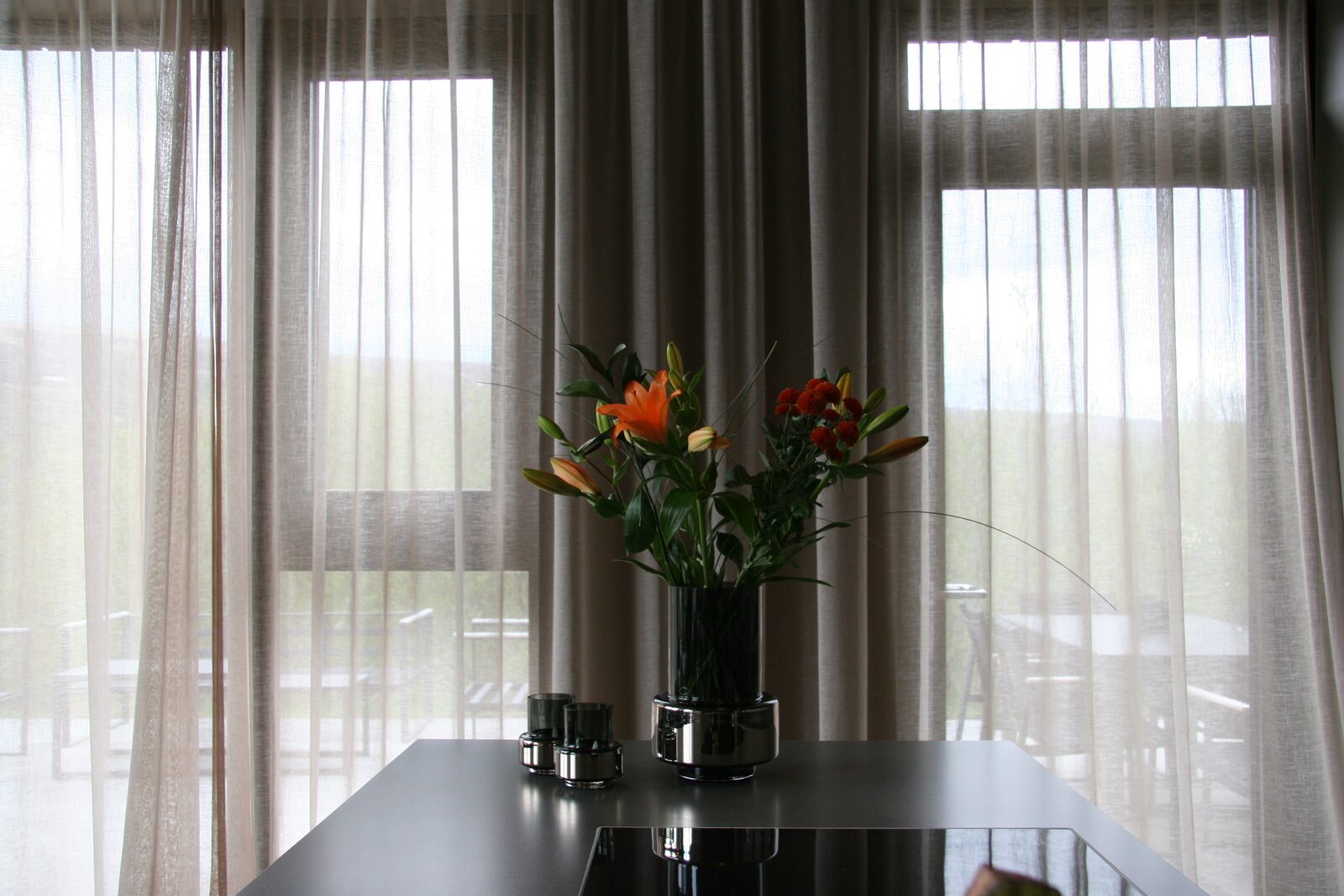 The dining table with orange flowers beside tall windows and sheer curtains.