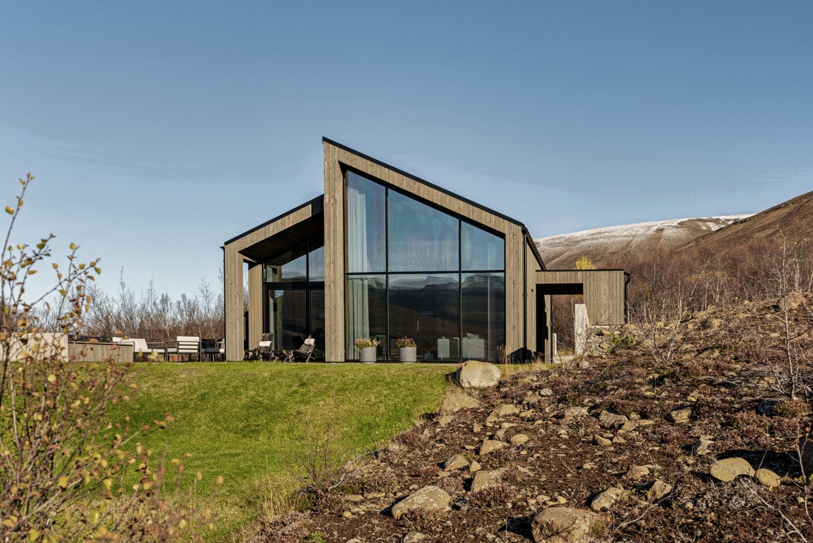 The glass gable and terrace of the house at Lundskógur seen from the grass bank under a clear blue sky, with a fell dusted with snow behind.