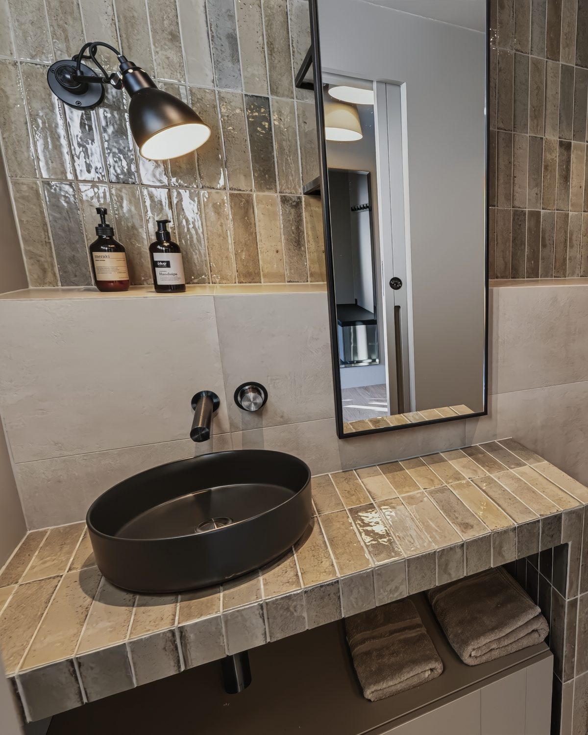 The extra WC at Lundskógur, a black basin on a tiled vanity under a wall light and a tall mirror, with toiletries on the shelf behind.