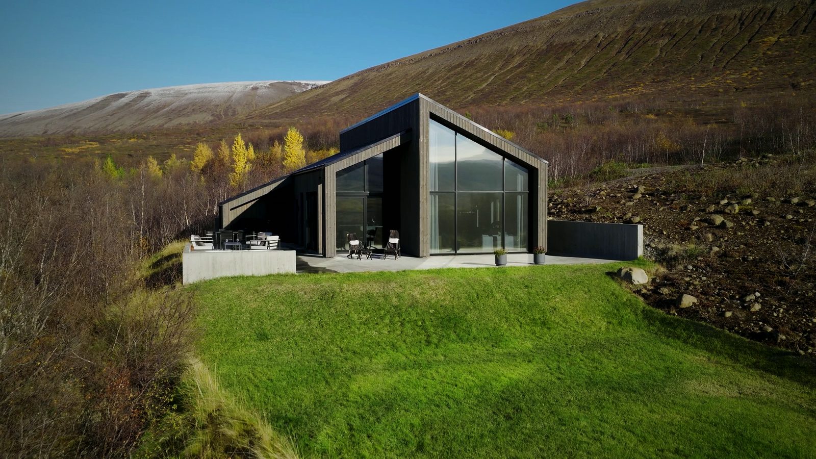 The glass gable and terrace of the house at Lundskógur in autumn sun, with green grass in front and a fell dusted with snow behind.