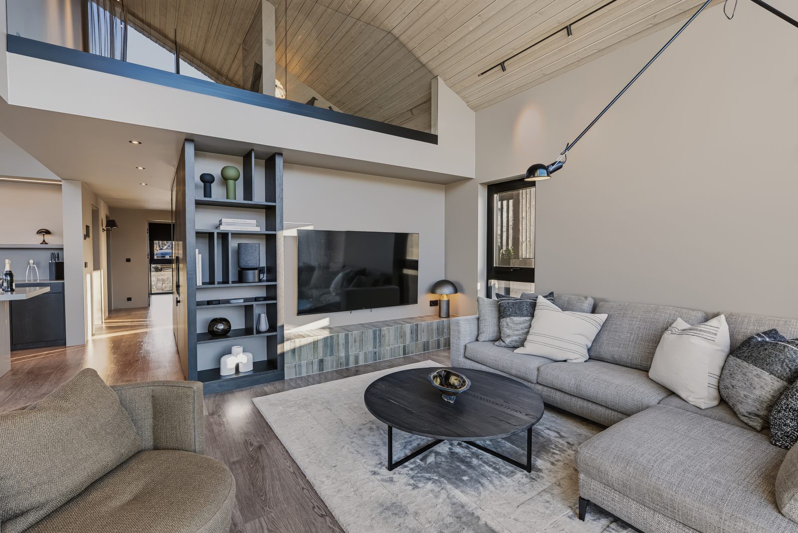 The living area at Lundskógur from the sofa, with the TV wall, open shelving and the upstairs balcony under the high timber ceiling.