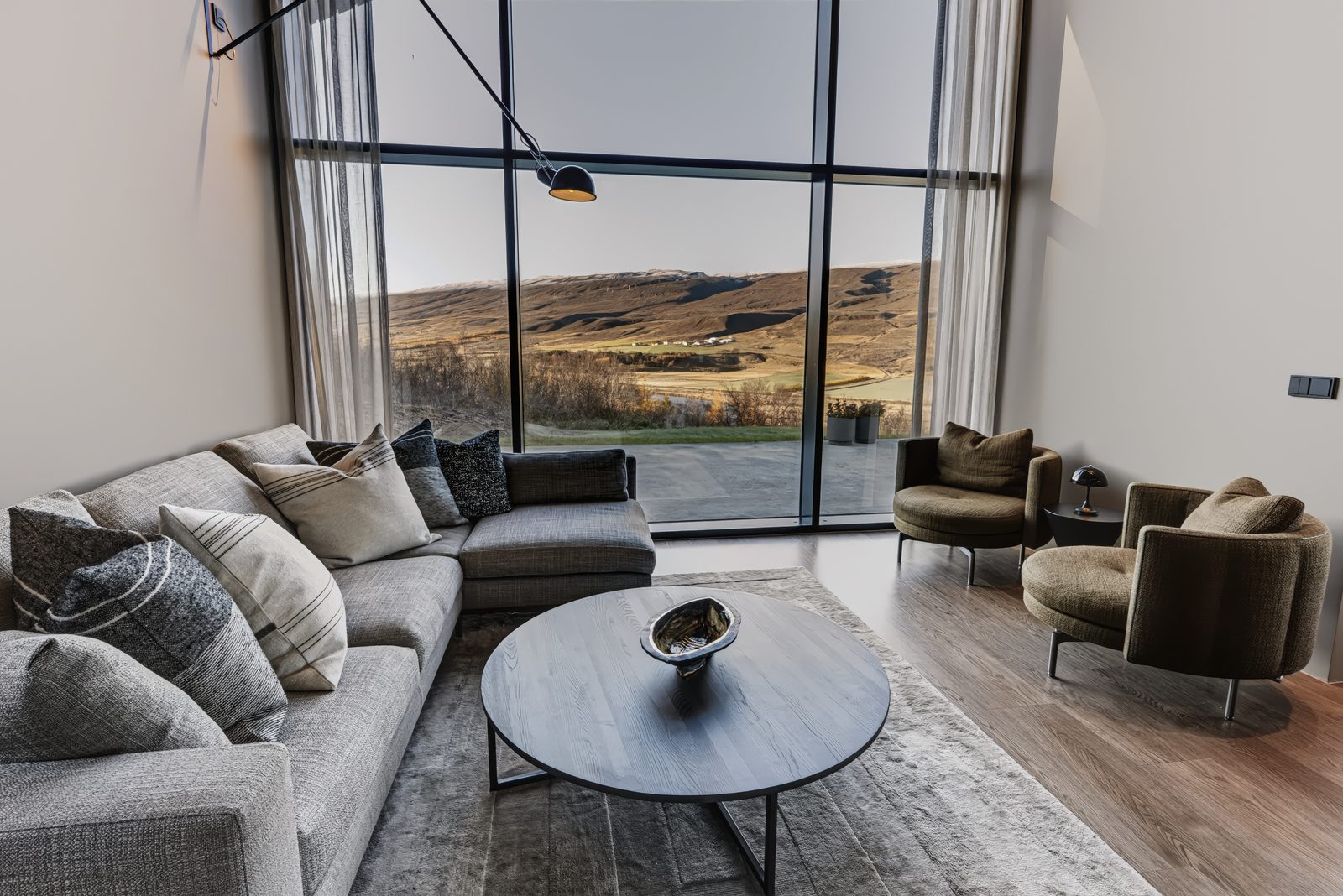 The living area at Lundskógur, with a grey sectional sofa, two olive armchairs and a round table in front of the full-height glass wall and the valley.
