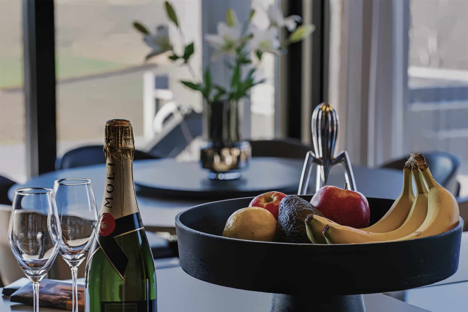 A bottle of champagne, two glasses and a bowl of fruit on the dining table, with flowers behind.