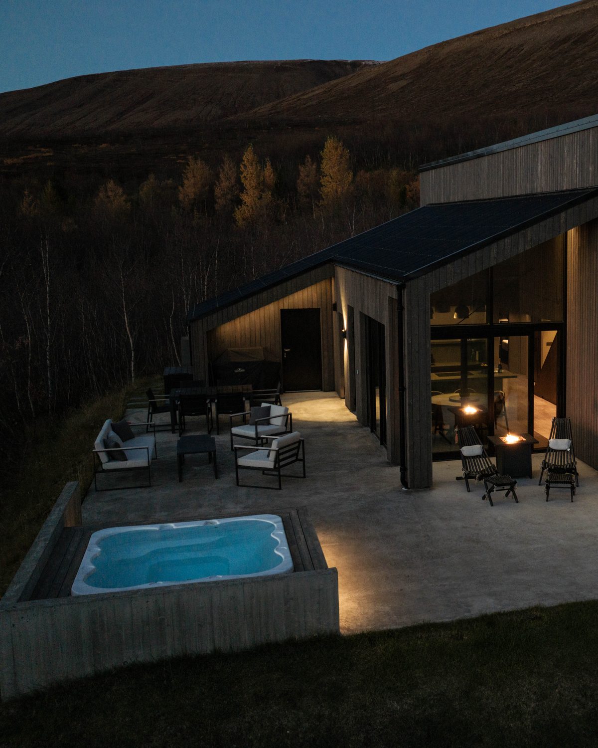 The terrace at Lundskógur at dusk from above, with the hot tub lit blue, the lit glass gable and the hillside behind.