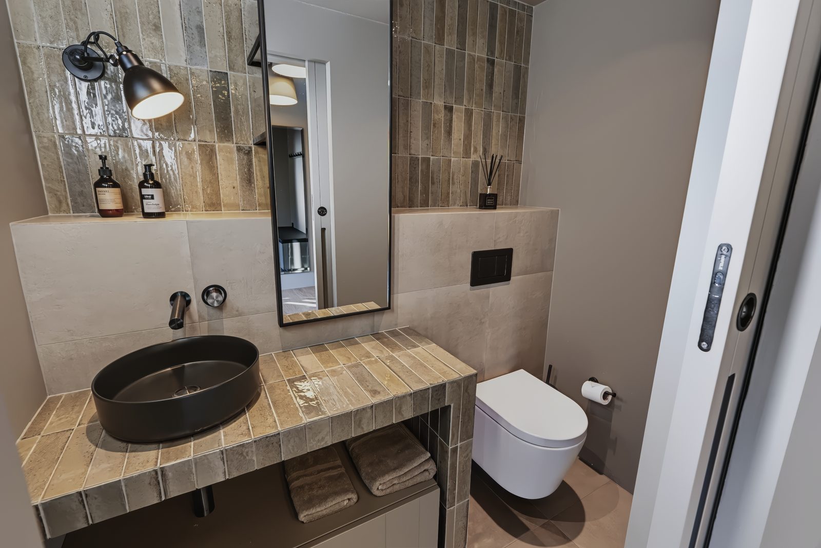The extra WC at Lundskógur, a black basin on a tiled vanity under a wall light and a tall mirror, with a wall-hung toilet beside it and a sliding door at the edge.
